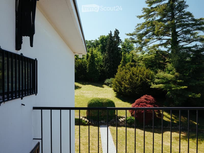 Balcony overlooking a well-maintained garden with various trees and shrubs, featuring a black metal railing and a view of the house's exterior wall.