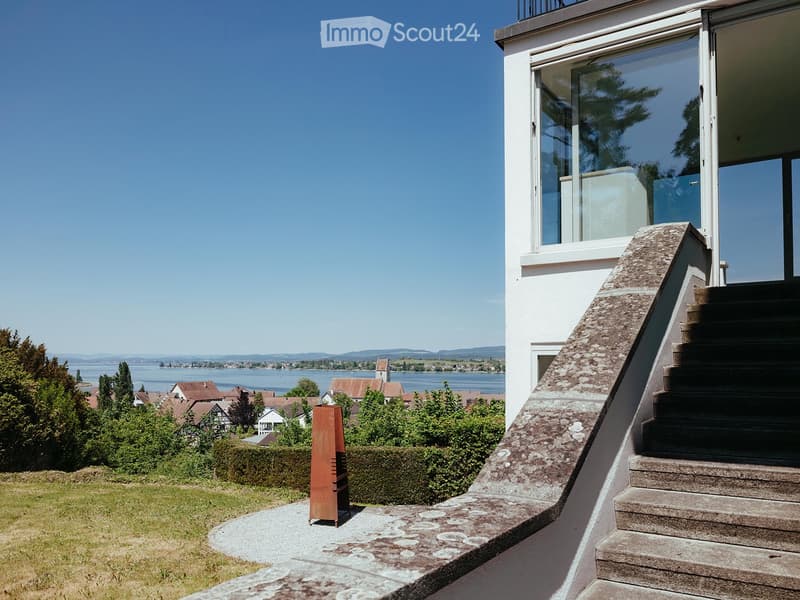 Garden area with a view of a lake and distant hills, featuring a stone staircase leading up to a modern house with large windows.