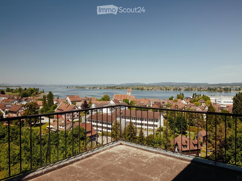 Balcony overlooking a town with houses, trees, and a lake in the background under a clear blue sky.