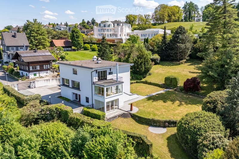 Aerial view of a modern white house with a balcony, surrounded by a well-maintained lawn and trees, with neighboring houses and a scenic hillside in the background.