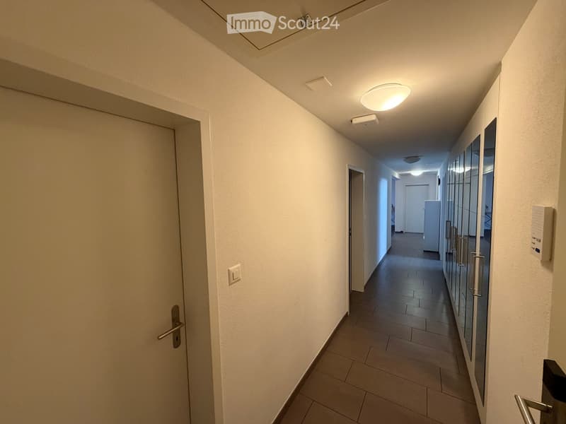 Clean, modern hallway with white walls, tiled floor, and recessed lighting. Features multiple doors and a glass-paneled wall on one side.
