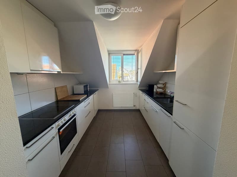 Modern kitchen with white cabinets, dark countertops, and built-in appliances including an oven and microwave. Features a window with blinds and a radiator below.