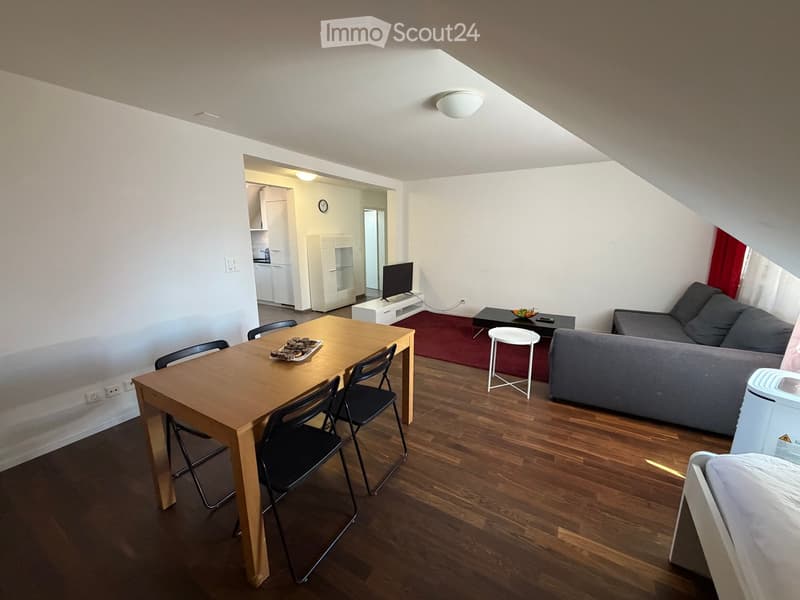 A modern, well-lit living space with a grey sofa, wooden dining table, black chairs, a small TV, and a white stool. The room features wooden flooring and white walls, with a small kitchen area visible in the background.