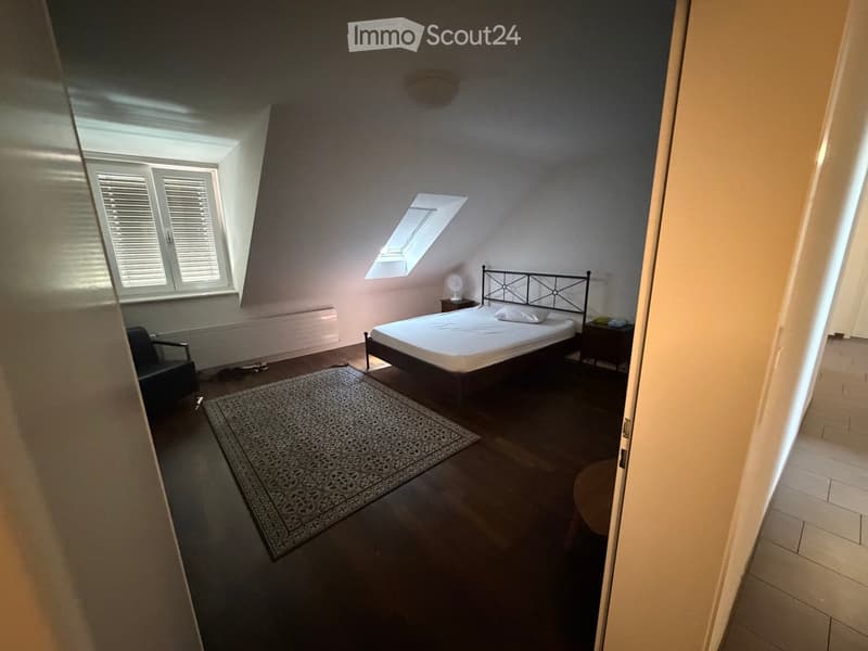 Bedroom with a bed, chair, small table, and fan. The room has wooden flooring, a patterned rug, and large windows with blinds.