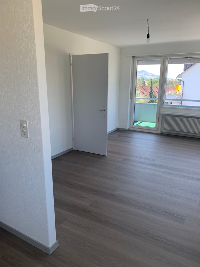Empty room with wooden flooring, white walls, a closed door, and a large window with a view of the outside area.