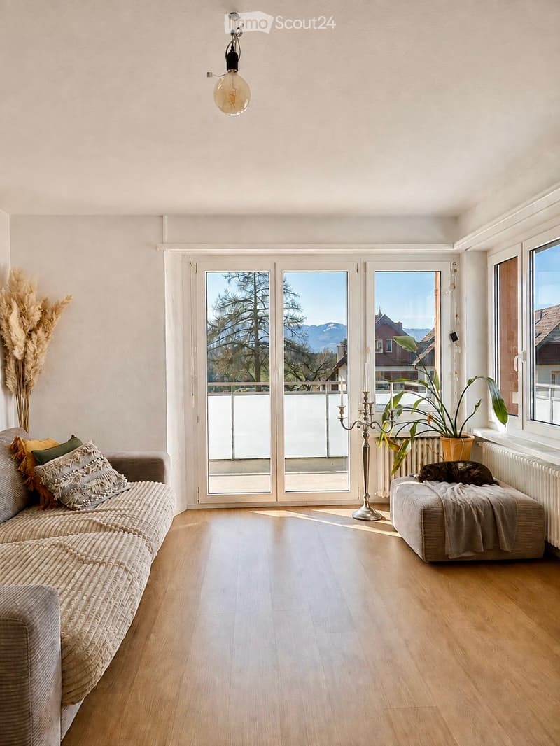 Bright living room with wooden flooring, large windows, and a view of the mountains. Features a woven sofa with pillows, an ottoman with a blanket, a candle holder, and a potted plant.