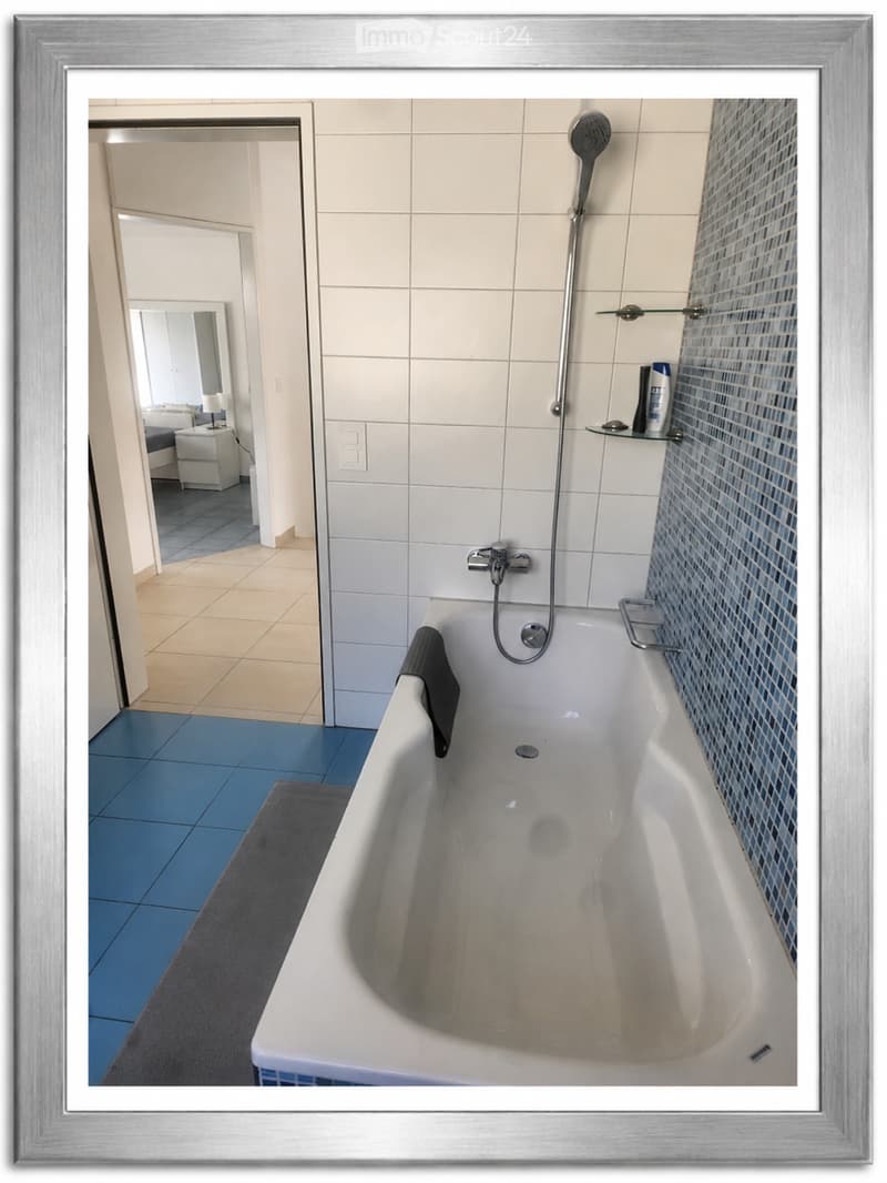 Modern bathroom with a white bathtub, glass shelf, and showerhead. Blue tiles on the floor and a grey towel on the side of the tub.