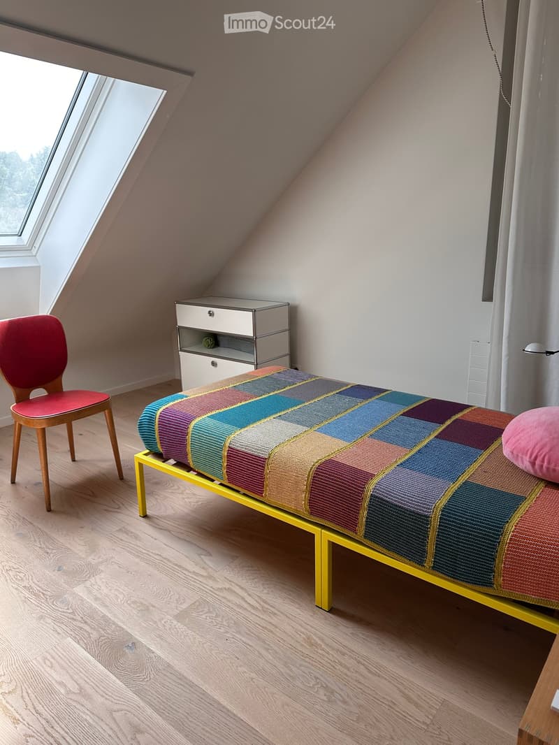 Modern bedroom with a colorful checkered bedspread, a wooden chair, and a sleek nightstand. The room features wooden flooring and large windows with a view.