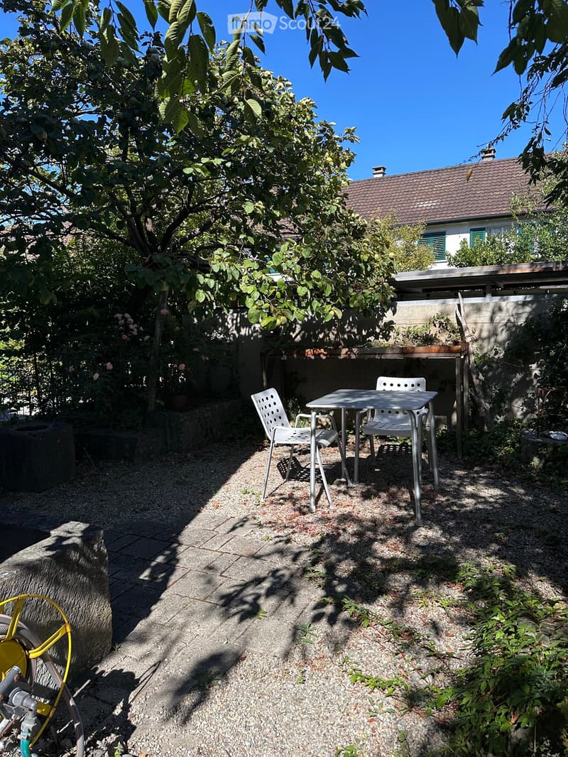 A small outdoor seating area with a table and two chairs, surrounded by greenery and a house in the background.