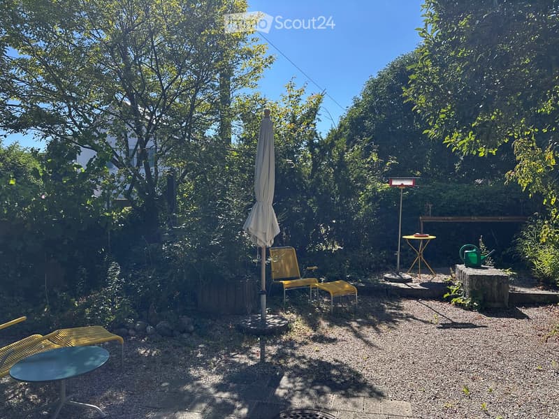 A small, well-maintained garden with gravel ground, surrounded by lush greenery. Features include a yellow lounge chair, a small round table, a closed umbrella, and a green watering can on a stone platform.