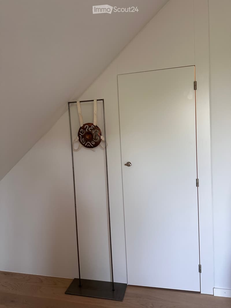 An unfurnished room with a wooden floor, white walls, and a white door. A tall, modern metal stand with a decorative circular object is placed near the door.
