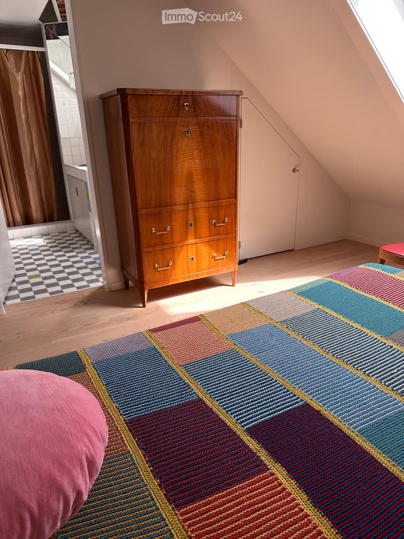 A cozy attic bedroom with wooden floors, a large wooden dresser, a colorful geometric rug, and a pink round cushion on the floor.