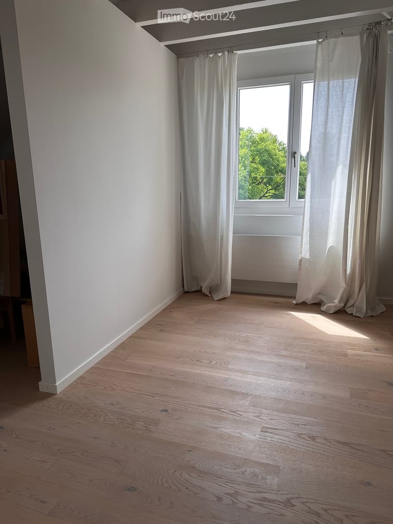 Empty room with light wooden flooring, white walls, and large windows with white curtains allowing natural light to enter.