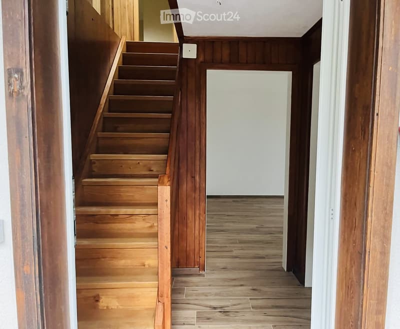 Wooden staircase leading upwards, wooden floor, white walls, open doorway to another room.