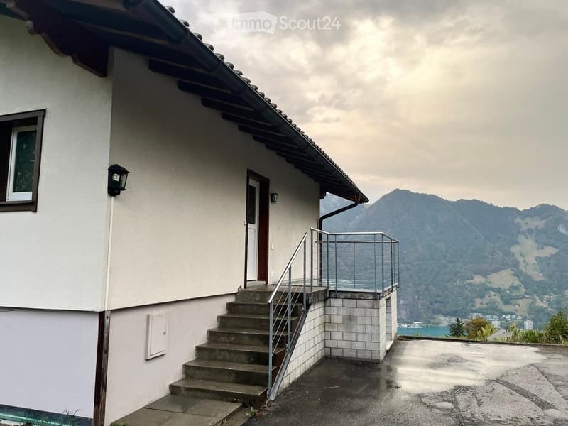 A two-story house with a white exterior, a covered entrance, and a staircase leading to a balcony overlooking a scenic mountain and lake view.