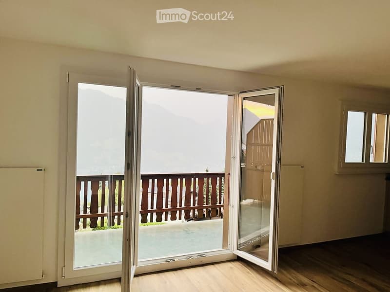 Open sliding doors lead to a wooden balcony with a railing, overlooking a scenic outdoor area.