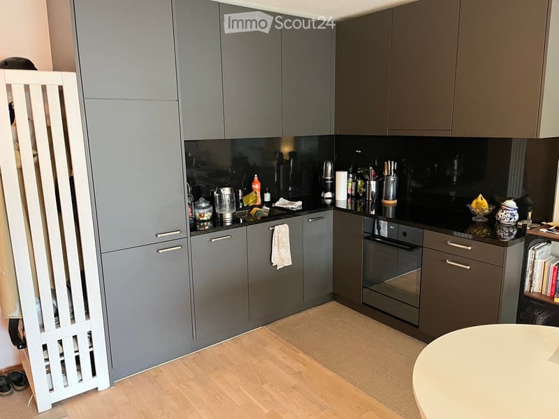 Modern kitchen with grey cabinets, stainless steel appliances, and a black countertop. Includes an oven, refrigerator, blender, and various kitchen items on the counter.