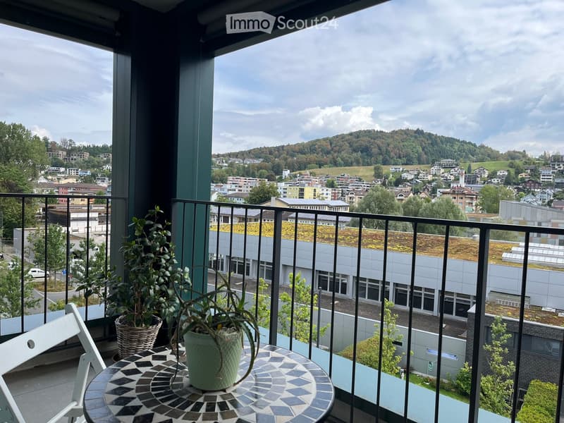 Balcony with a view of a cityscape and hills, featuring a small table with two potted plants and a white chair.