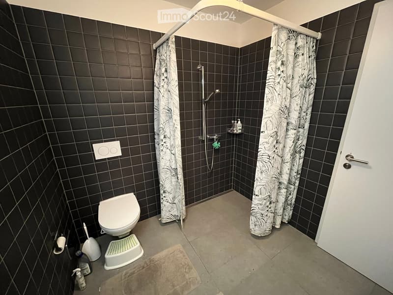 Modern bathroom with dark tiled walls, a toilet, a shower area with a curtain, and various cleaning supplies and a towel on the floor.