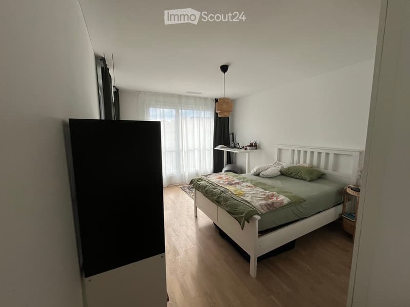 A well-lit bedroom with a wooden floor, a white bed with green bedding, a nightstand, a small table with a chair, and a hanging lamp.