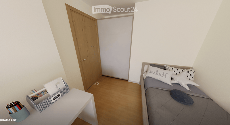 A small, modern bedroom with a single bed, a laptop on a small table, and a cup with pencils nearby. The room features light-colored walls and wooden flooring.