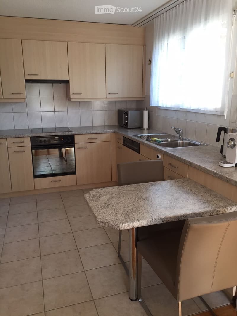Modern kitchen with light wood cabinets, granite countertops, stainless steel appliances, and a small dining area with a chair and table.