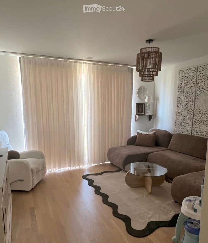 Modern living room with a large window covered by beige curtains, a brown sectional sofa, a glass coffee table, a white armchair, and a decorative wall piece.