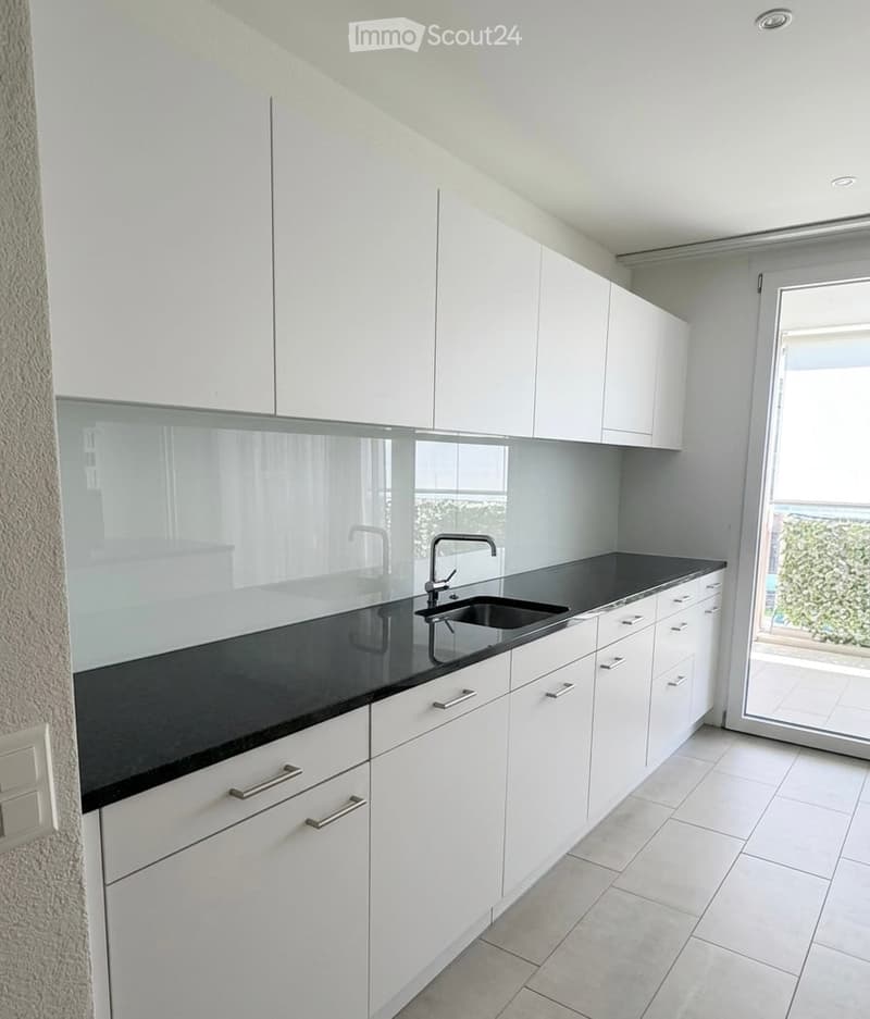 Modern kitchen with white cabinets, black countertop, and a sink with a modern faucet. The space is bright with large windows and a sliding glass door leading to a balcony.