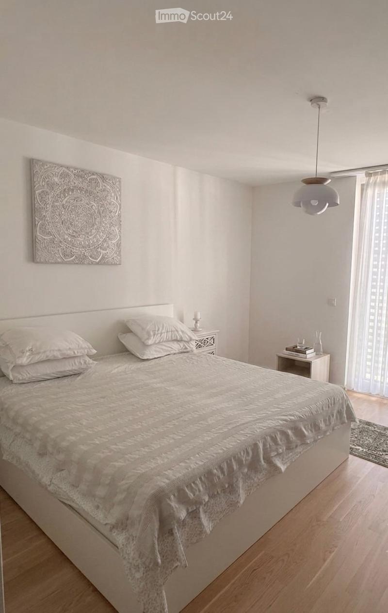 Modern bedroom with a large bed, white bedding, nightstand, lamp, decorative wall art, and a vase on a small table near the window.