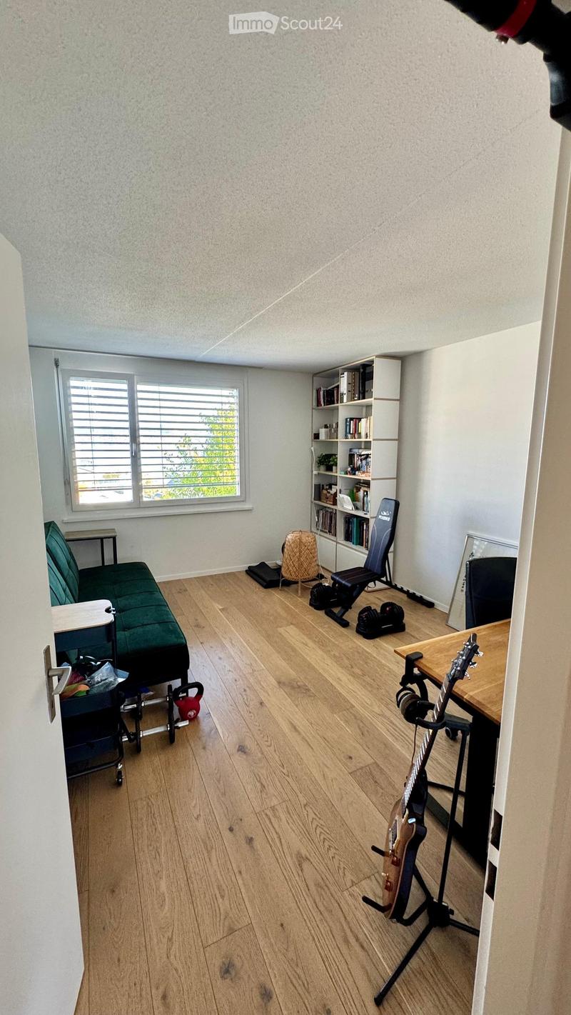 A multi-purpose room with wooden flooring, a green sofa, a guitar on a stand, a bookshelf, and exercise equipment.