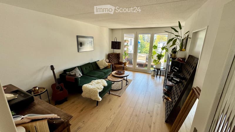 Modern living room with a green sectional sofa, wooden coffee table, guitar, and large windows leading to a balcony.