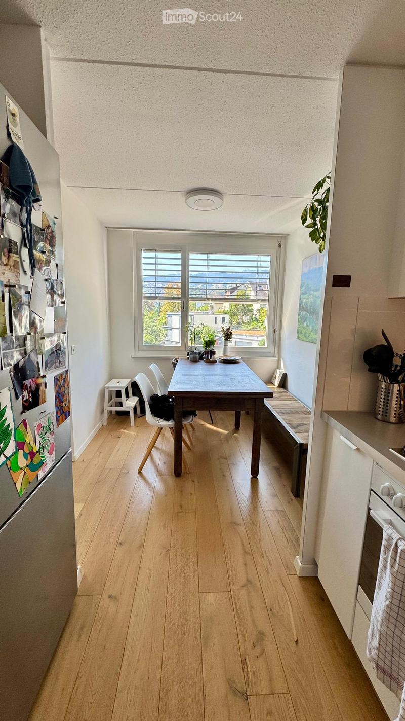 Bright kitchen with wooden flooring, a dining table with chairs, a small stool, a refrigerator with photos, and a window with a view of the city.