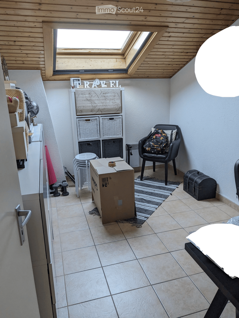 A small, furnished room with a skylight, tiled floor, and various items including a chair with a backpack, a storage unit with baskets, a globe, a lamp, and a small table.