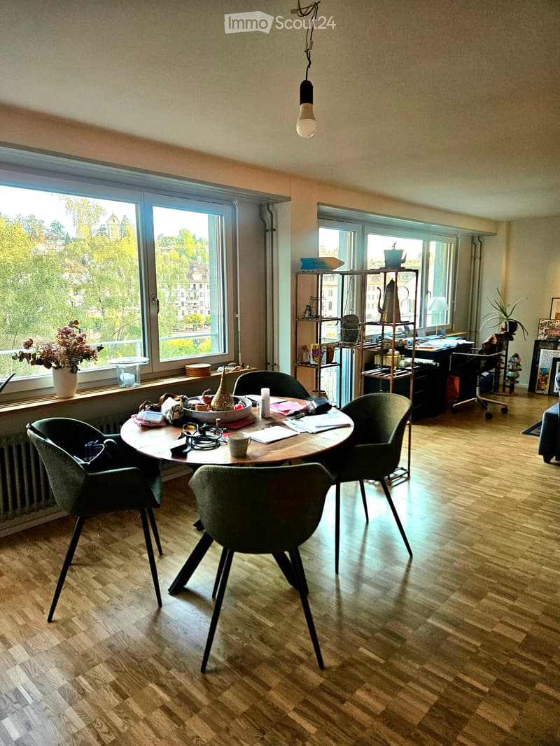 Bright room with wooden flooring, large windows, a round table with four chairs, a metal shelving unit, and decorative items on the windowsill.