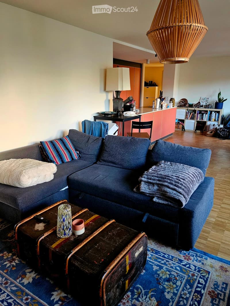 A modern living room with a blue sectional sofa, a wooden coffee table, a large hanging lamp, and a small kitchen area visible in the background.