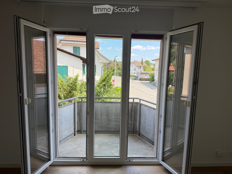 Open sliding doors leading to a balcony with a concrete floor and metal railing, overlooking a residential area with houses and greenery.
