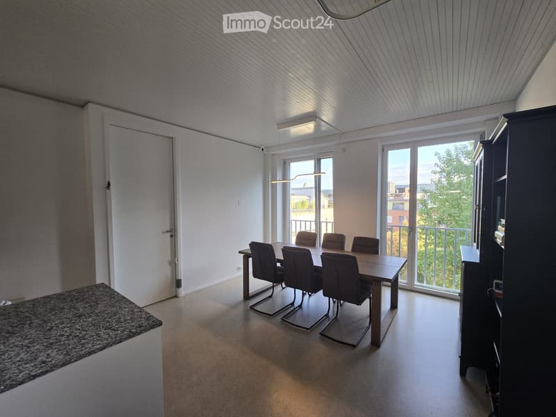 Bright dining area with a wooden table and six chairs, large windows and sliding doors leading to a balcony, modern lighting fixtures, and a dark cabinet.