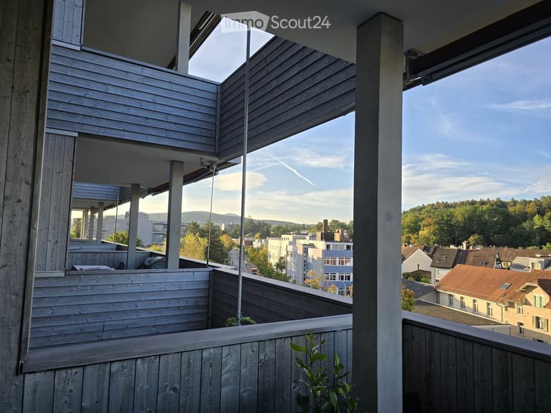 Wooden balcony with open sides, overlooking a cityscape with buildings and trees.