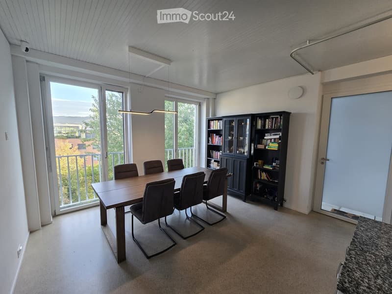 Bright room with a wooden dining table and six chairs, a large bookshelf, and a cabinet filled with books and decorative items. Large windows and sliding doors lead to a balcony with a view of greenery.