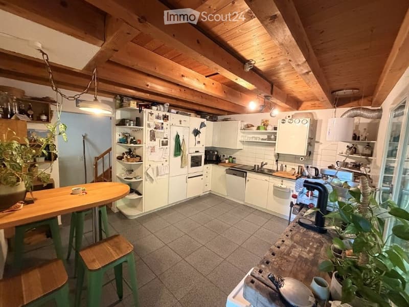 Cozy kitchen with wooden ceiling beams, white cabinets, and a central island. Includes a refrigerator, oven, dishwasher, sink, and various kitchen appliances. There is a wooden table with green stools and potted plants on the counter.