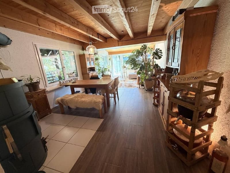 Spacious, well-lit room with wooden beams, hardwood flooring, and large windows. Features a dining table with chairs, a fur-covered bench, a wooden cabinet, a wooden shelf, and several potted plants. A wooden stove is visible on the left side.