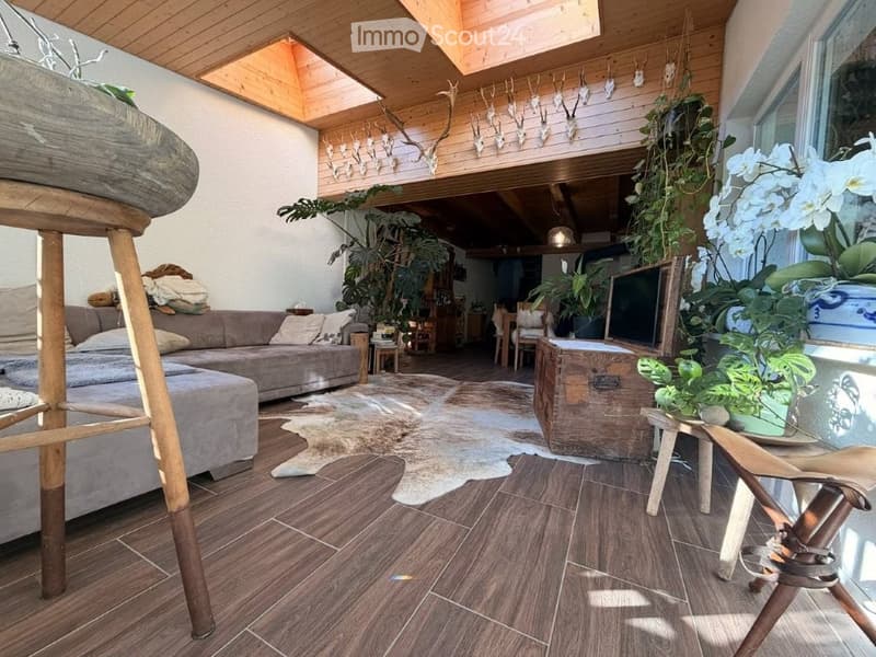 A modern living room with a grey sectional sofa, ottoman, wooden floor, large windows, and various plants. A television is placed on a wooden stand, and there are decorative items and a rustic wooden chest in the space.