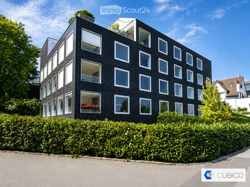 Modern black brick building with multiple large windows and balconies, surrounded by greenery and a hedge.