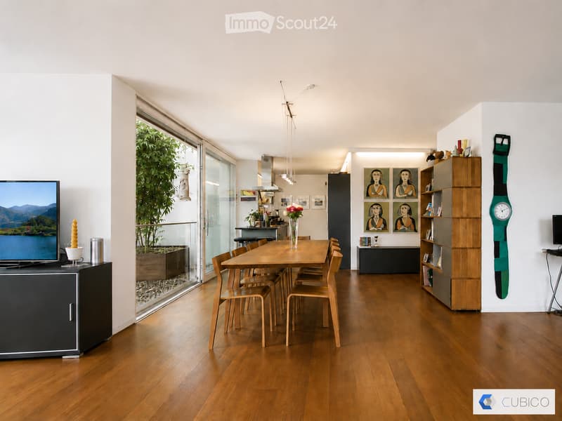 A modern, open-plan dining area with a wooden dining table and chairs, a television on a cabinet, a large window with a view, and a shelf with decorative items.