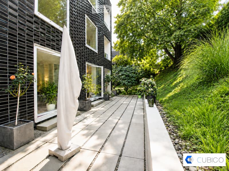 Modern outdoor patio area with a sleek, black-tiled house, large windows, and a well-maintained garden with various plants and trees. Features include a tall white parasol, stone pathway, and decorative planters.