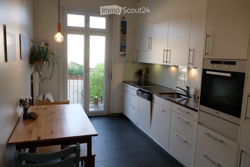 Modern kitchen with white cabinets, stainless steel appliances, a wooden dining table, and a hanging light fixture. A sliding glass door leads to a balcony with outdoor plants.