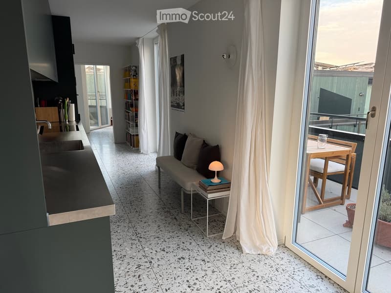 Modern open-plan living and dining area with a sofa, small table, and a kitchen counter. Large windows lead to a balcony with a small table and chairs.