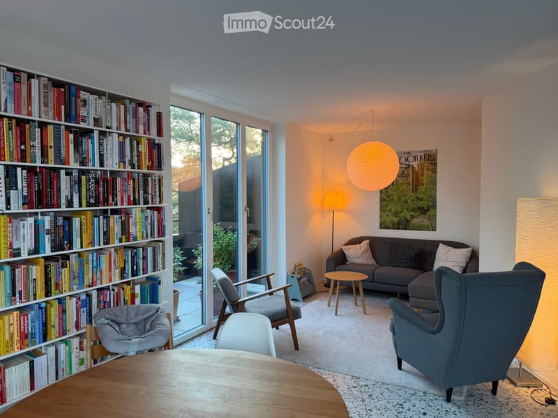 Modern living room with large windows, a bookshelf filled with books, a grey sofa, wooden chairs, a small round table, and a hanging lamp.