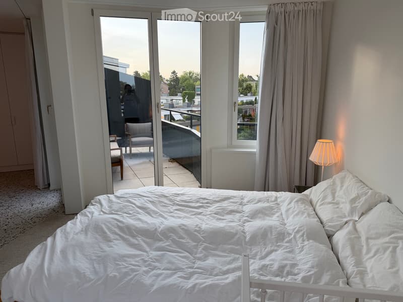 Bright bedroom with a white bed, nightstand, and lamp. Large sliding glass doors open to a balcony with outdoor seating.