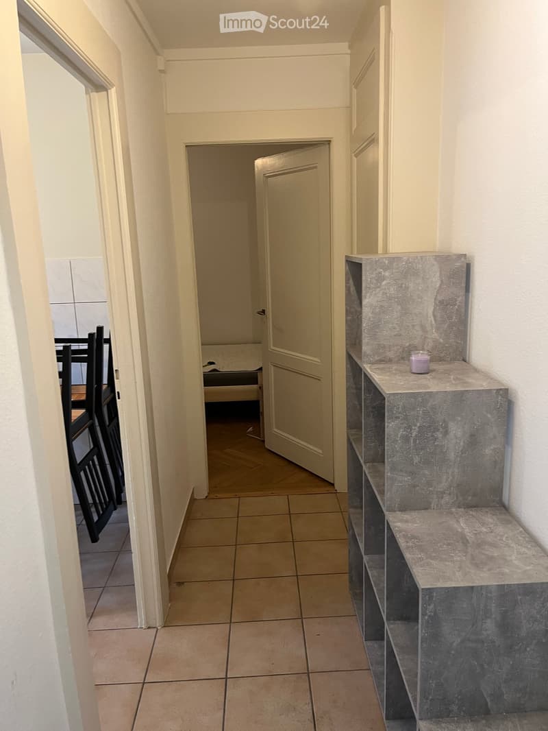 A narrow hallway with tiled flooring, white walls, and a marble bench on the right side. A door leads to another room, and a glimpse of a dining area is visible on the left.